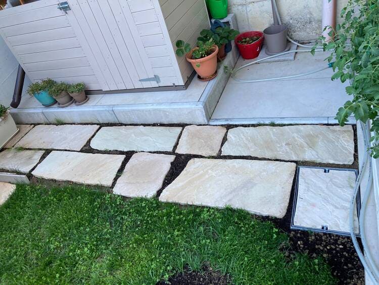 Vista dall’alto di un sentiero da giardino creato da Zini Dino con grandi lastre di pietra naturale irregolare (tinte sabbia e grigio chiaro) incassate nel terreno e circondate da erba verde. Il sentiero conduce a un’area patio pavimentata in grigio con vasi di piante e un armadietto di legno. Sulla destra, una lastra di copertura per tombino in pietra.
